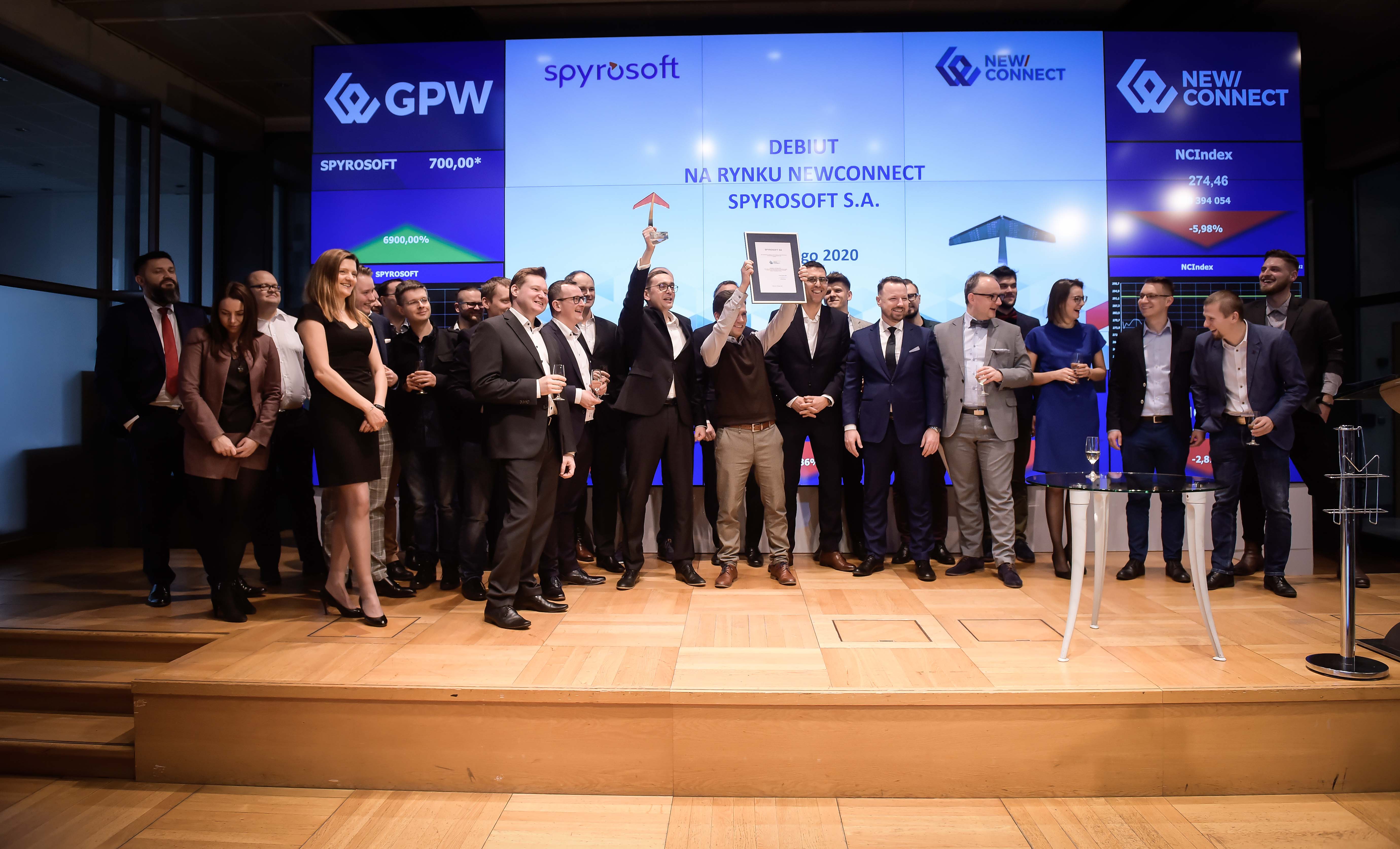 Spyrosoft team celebrating their debut on the NewConnect stock market, holding documents on stage at the GPW exchange.