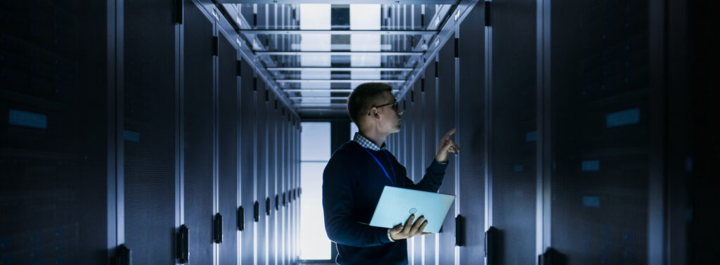 Male IT Engineer Works on a Laptop in front of Server Cabinet at a Big Data Center. Rows of Rack Servers are Seen.