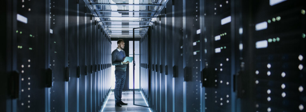 IT Technician Works on Laptop in Big Data Center full of Rack Servers. He Runs Diagnostics and Maintenance, Sets System Up.