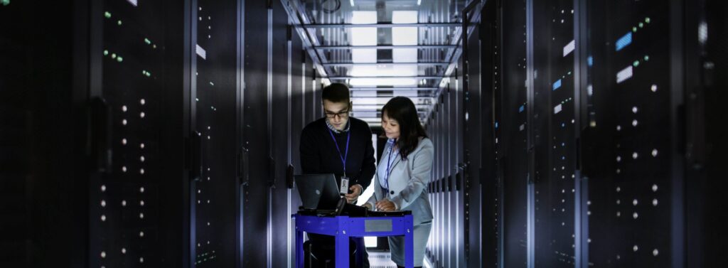 A view of a large IT server room. In the middle stand a man and a woman, employees, analyzing data on their laptop.
