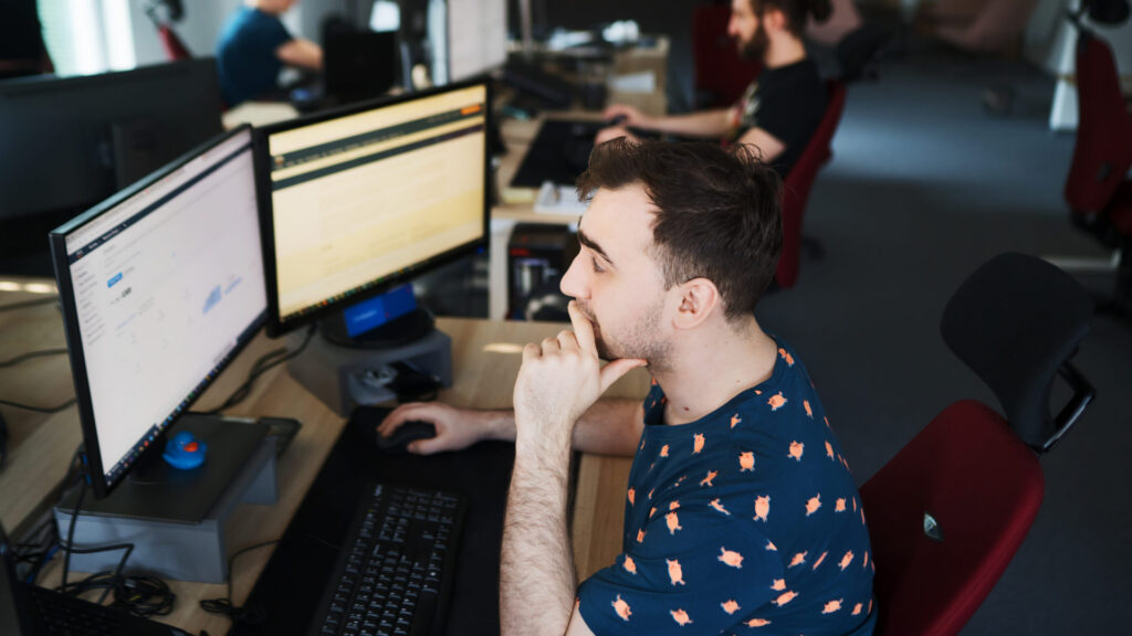 kupiony programista z polskiej firmy IT pracuje w nowoczesnym biurze, analizując dane na dwóch monitorach, co ilustruje wysoką jakość usług i jeden z 8 powodów, by rozpocząć współpracę.