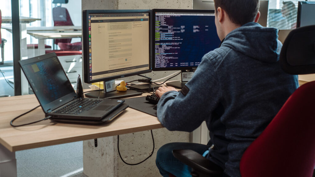 A data scientist works at a desk with a laptop and two monitors, writing code and analyzing data to build a customer lifetime value (CLV) prediction model