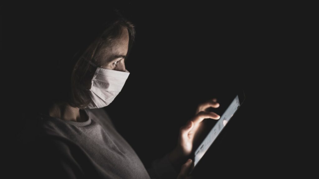 A person wearing a face mask in a dark room uses a tablet to access virtual healthcare, symbolizing the uncertainties and common myths surrounding the telemedicine sector