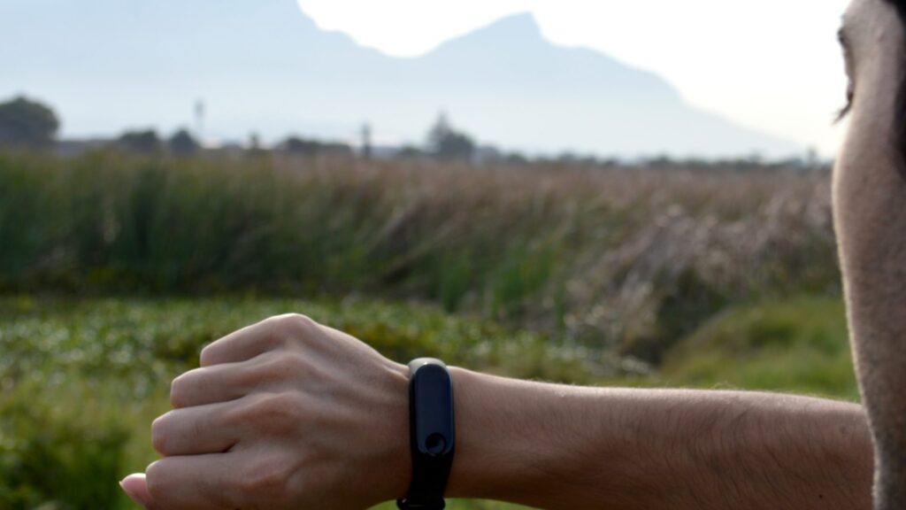 A person outdoors checking their MedBand wellness tracker on their wrist, monitoring their health data against a natural background of a field and mountains.