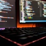 A developer's dual-monitor workstation displaying complex source code in a dark room, symbolizing the deep and essential work involved in software maintenance