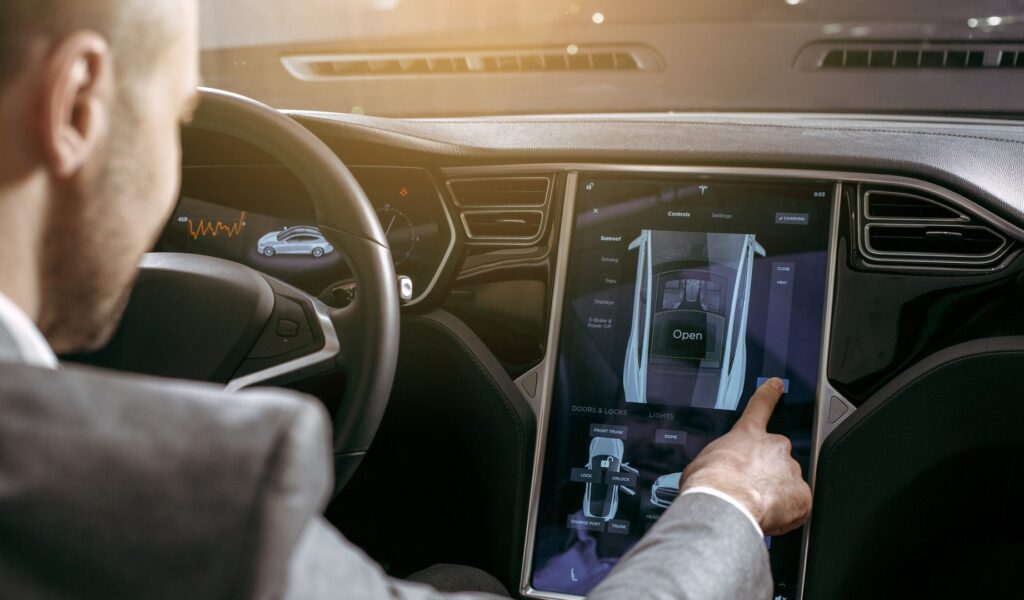 A person seated in the driver’s seat interacts with a large automotive touchscreen interface, selecting vehicle functions such as doors, locks, and climate features. The image represents modern in-car HMI systems that integrate haptic feedback to enhance user interaction, precision, and safety while operating digital controls inside a vehicle.