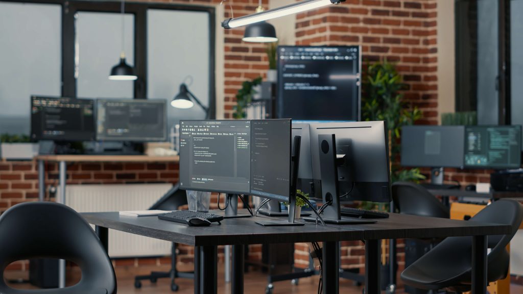 Developer workstation with dual monitors showing code in a modern open-plan office with exposed brick walls