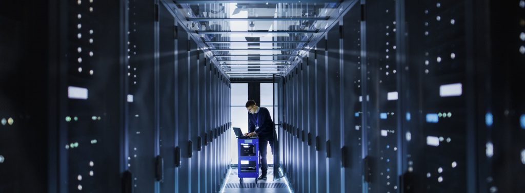 a view of a large server room, with a server room technician checking settings on a laptop at the end of the corridor