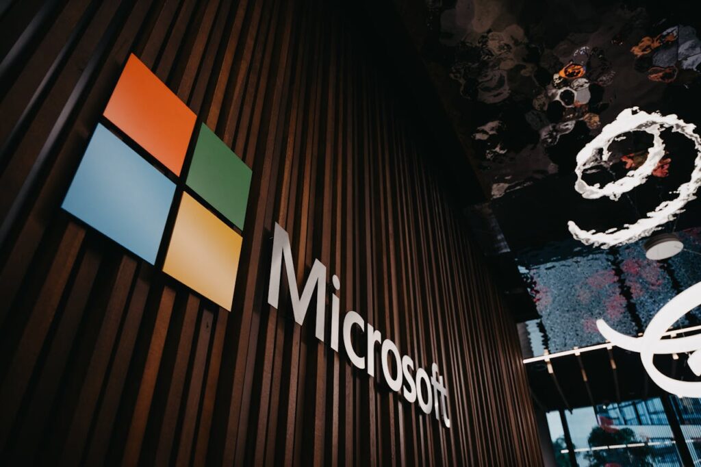 The Microsoft logo and wordmark mounted on a dark wooden slatted wall, photographed from a low angle. The four-colour Windows symbol in red, blue, green, and yellow is displayed alongside the white "Microsoft" lettering.
