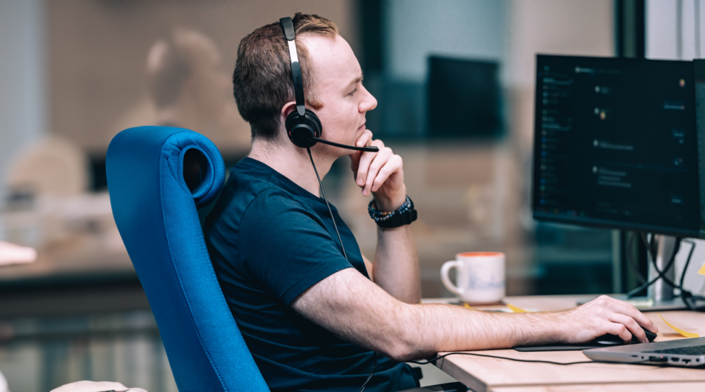 a developer sitting at his computer desk in the Spyrosoft office working on AWS Lambda project and thinking about the solution, lightly smiling and wearing headphones