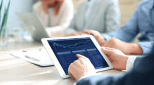 A business meeting, the main person is holding a tablet displaying stock market data.