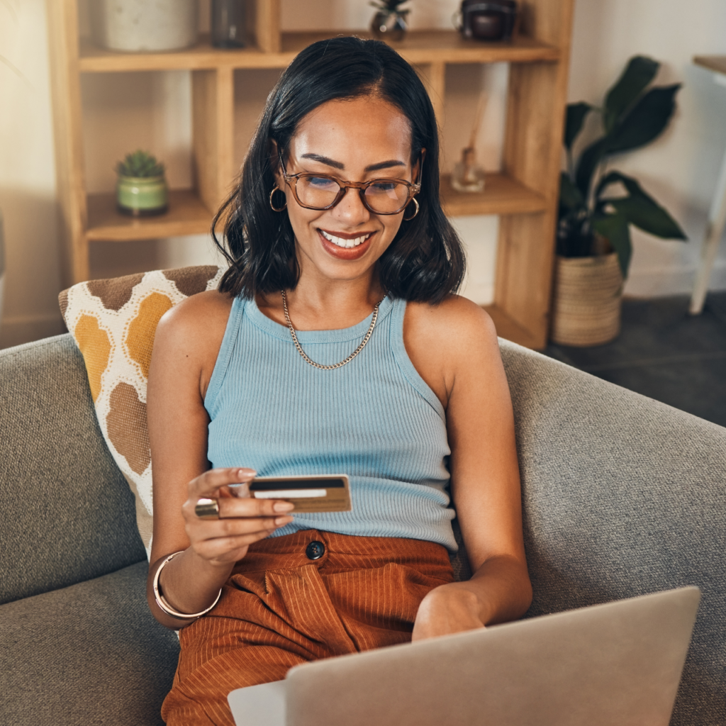 A smiling woman is sitting on a couch with a credit card in her hand and a laptop on her lap.