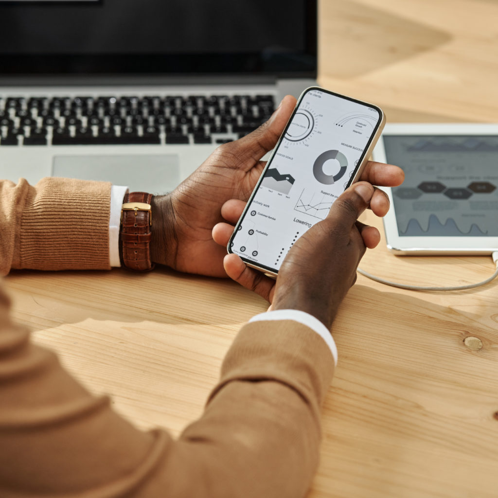 View from a person's perspective, a man holding a phone in his hand and analysing charts showing the results of his project; a laptop and tablet in the background.