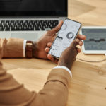View from a person's perspective, a man holding a phone in his hand and analysing charts showing the results of his project; a laptop and tablet in the background.