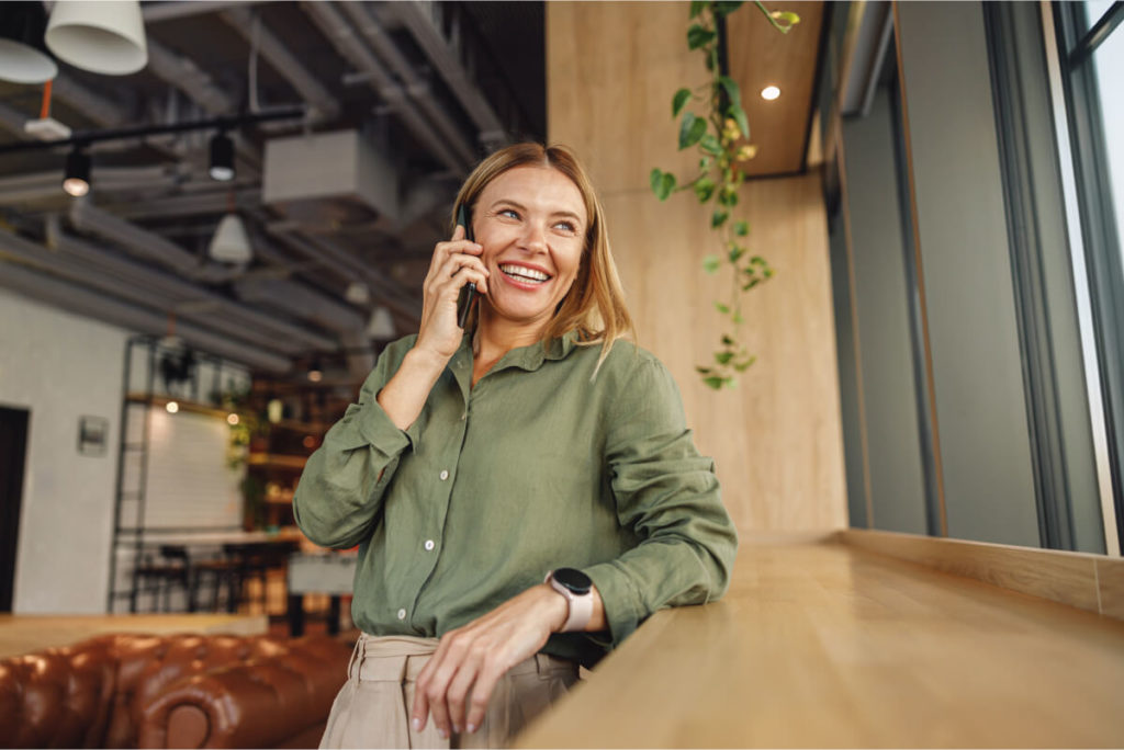 A smiling woman in a loft apartment, leaning against a windowsill, talking on the phone and looking out the window.
