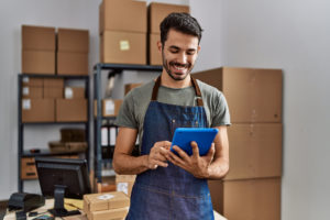 zdjęcie mężczyzny trzymającego tablet w dłoni i przeglądającego stronę sklepu internetowego e-commerce, stojącego w pomieszczeniu z paczkami przygotowanymi do wysyłki