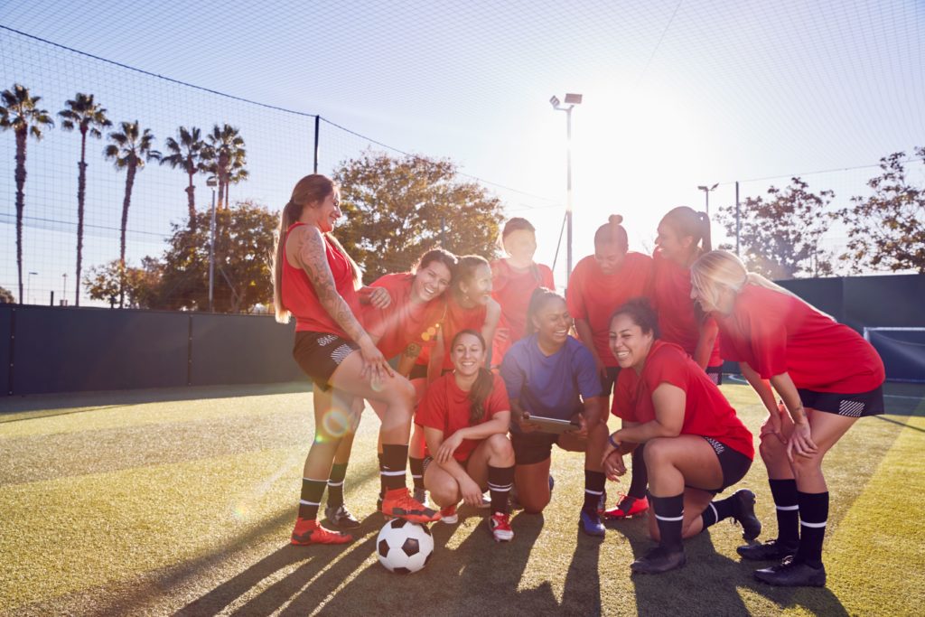 Sports streaming – women's football team smiling and celebrating together on outdoor training field with sunlight and net in background