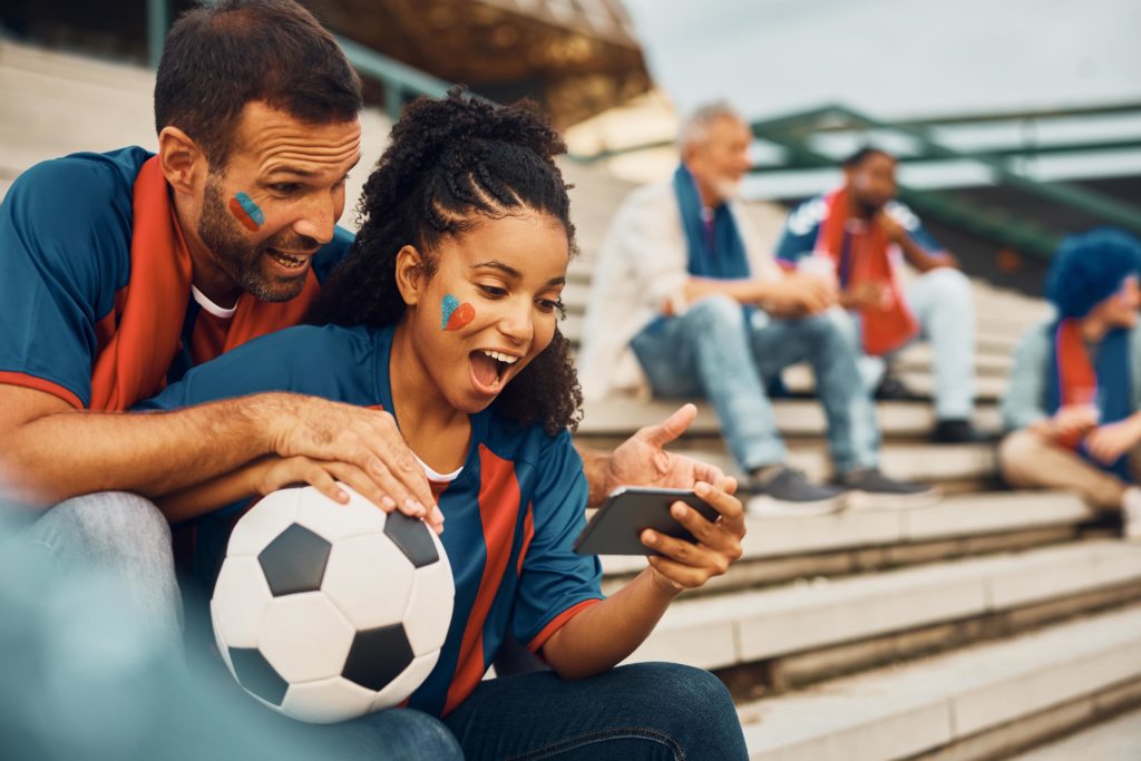 Sports streaming – football fans in jerseys watching match highlights on smartphone while sitting in stadium stands