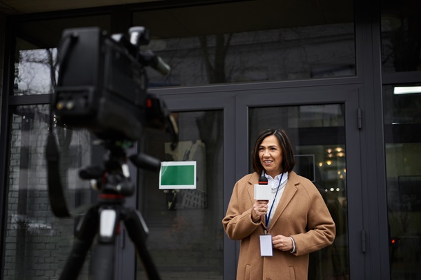 A multiracial female journalist in a beige coat actively shares news updates while holding a microphone outdoors, broadcasters