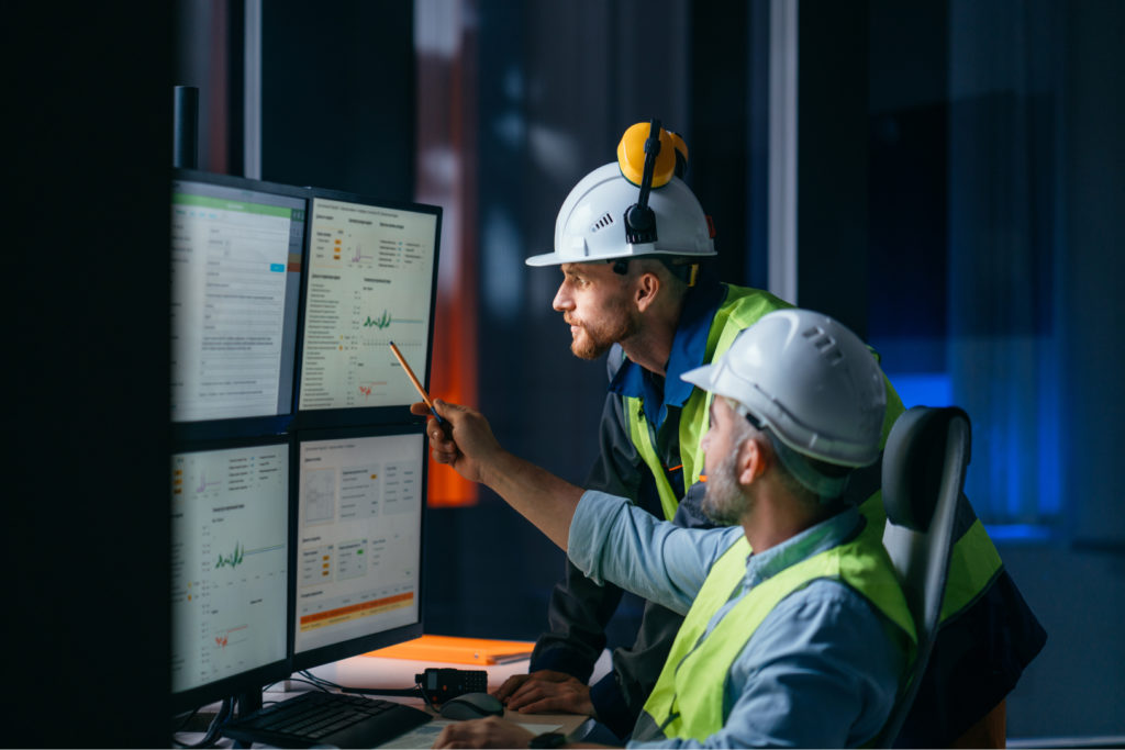 two men working in industry 4.0 wearing helmets looking and pointing at the screens with dashboards
