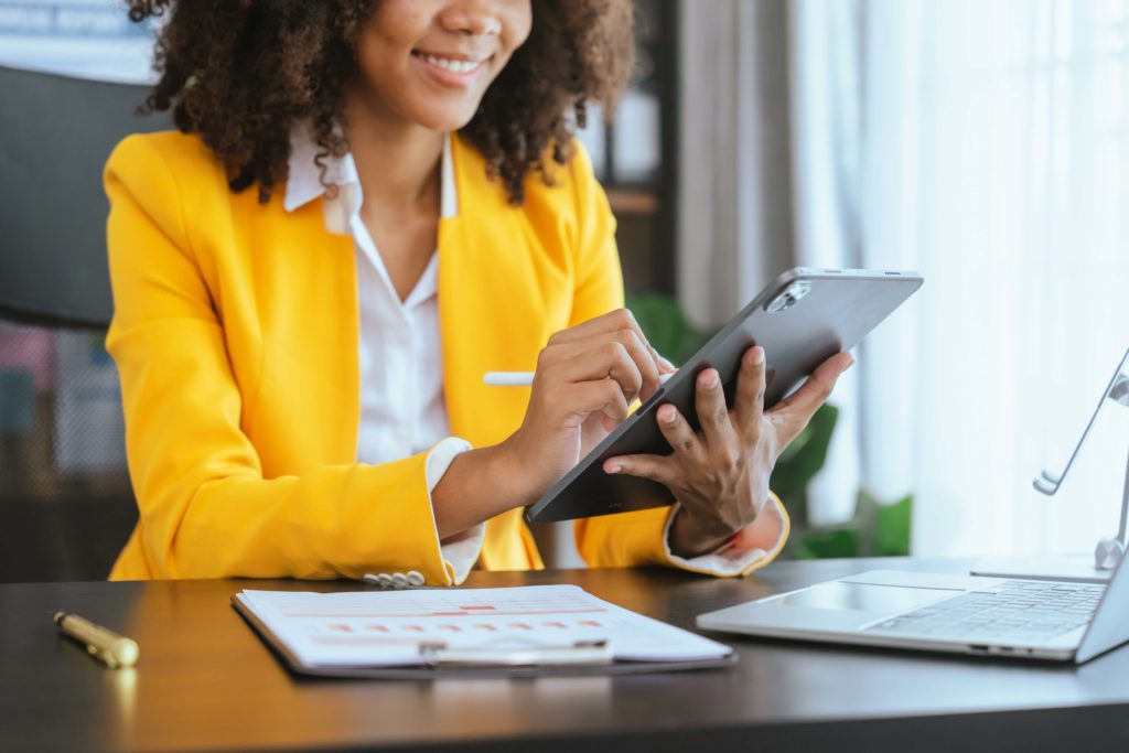 a woman in a yellow blazer sitting in the office smiling and clicking on a tablet