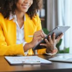 a woman in a yellow blazer sitting in the office smiling and clicking on a tablet