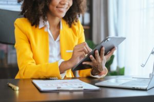 a woman in a yellow blazer sitting in the office smiling and clicking on a tablet