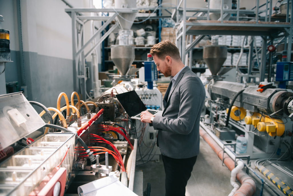 a manager inspects a machine in a manufacturing plant, holding a laptop