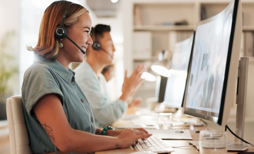 woman working in customer support sitting in an office at her desk in front of the computer screen wearing a headset and having a call with a client