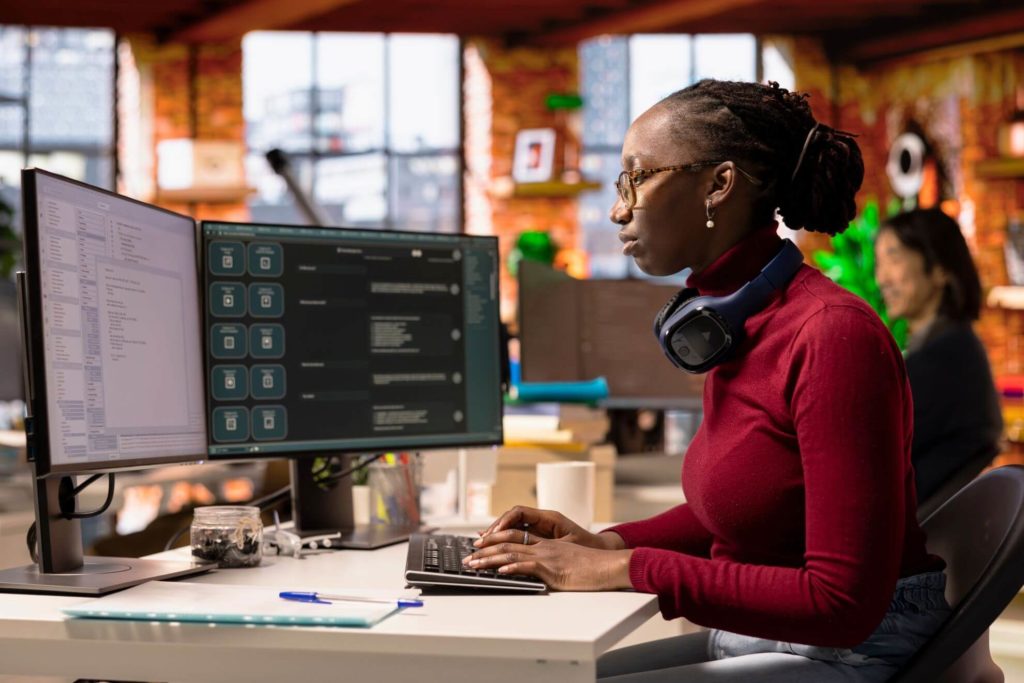 Software developer working at dual monitors in a modern office, coding with headphones around her neck.