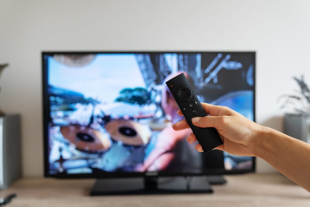 Hand holding a TV remote control pointed at a smart television with a blurred streaming interface on screen.