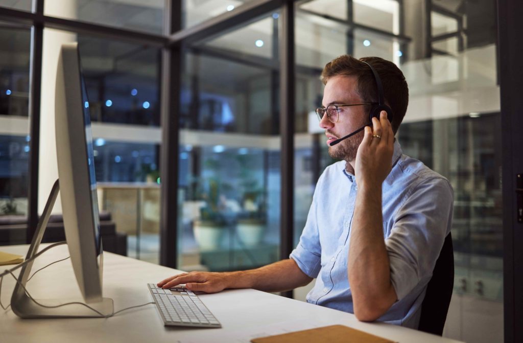 Man wearing headset and glasses working at desktop computer in modern glass office at night