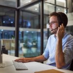 Man wearing headset and glasses working at desktop computer in modern glass office at night