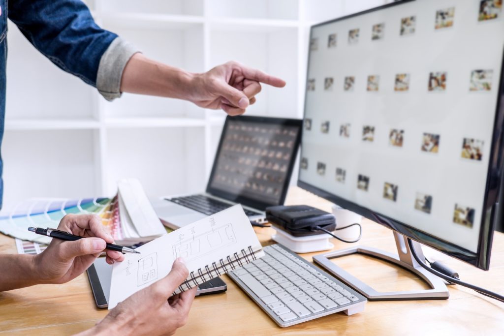 Two designers collaborating at a workstation. One person points at a large monitor displaying a grid of UI or photo thumbnails, while the other holds a pen and a spiral notebook with hand-drawn wireframe sketches. A laptop, graphics tablet, colour swatches, and a keyboard are visible on the desk.
