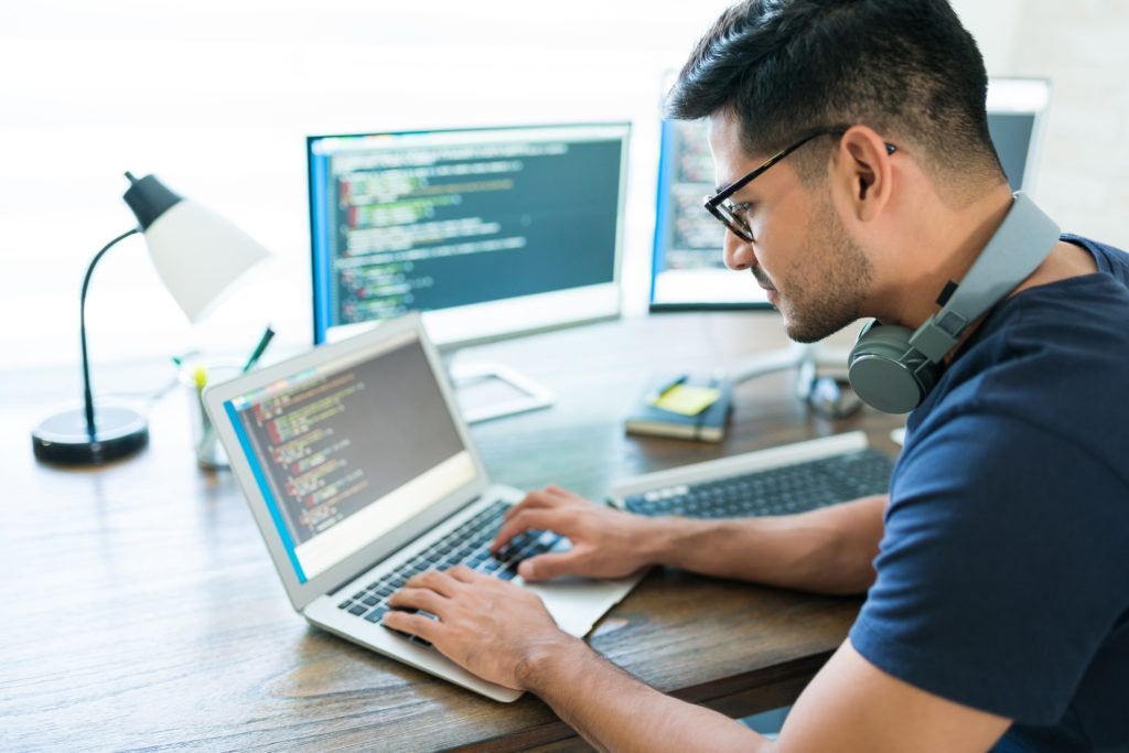 Developer working at a wooden desk, typing on a laptop with two additional monitors in the background, all displaying colourful code. He is wearing glasses, headphones around his neck, and a navy t-shirt.