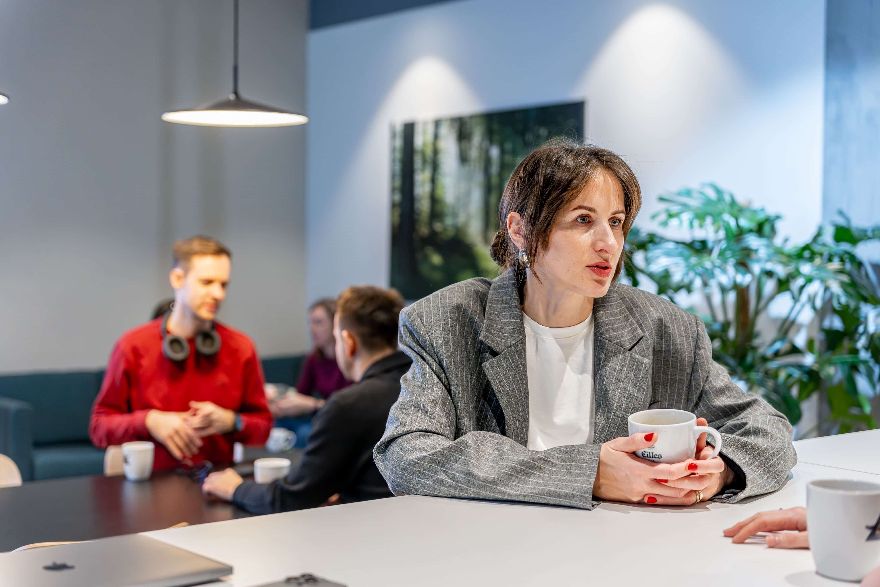 A woman sitting at the table with a cup in her hands is talking. There are a few people in the background talking.