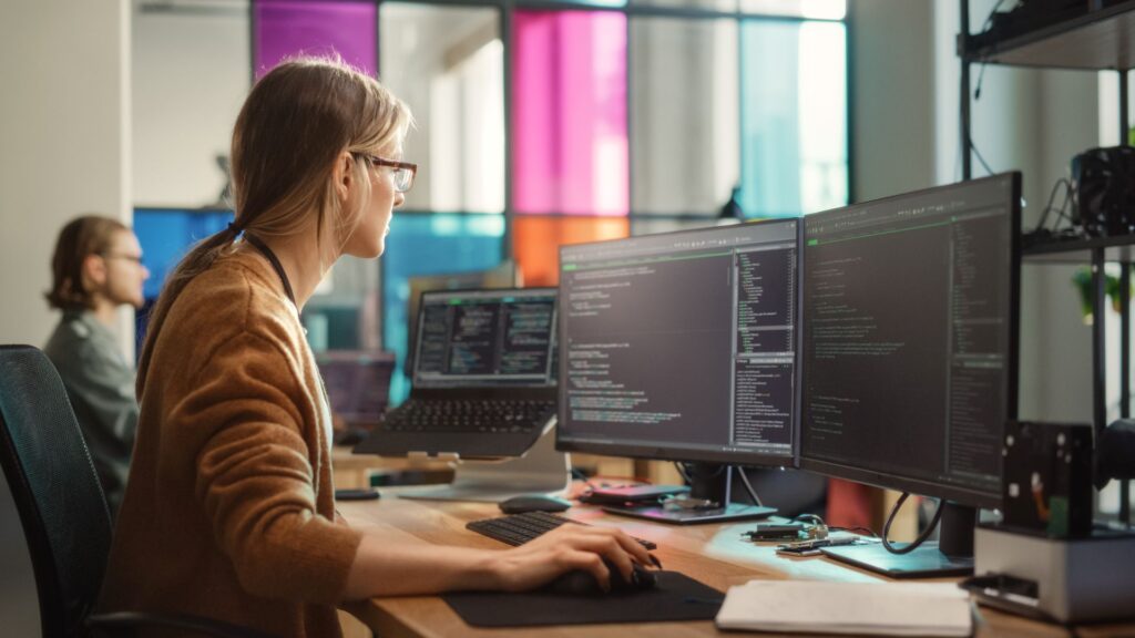 AI assisted software development. AI knowledge management. Female Software Engineer Writing Code on Desktop Computer with Multiple Screens Setup.