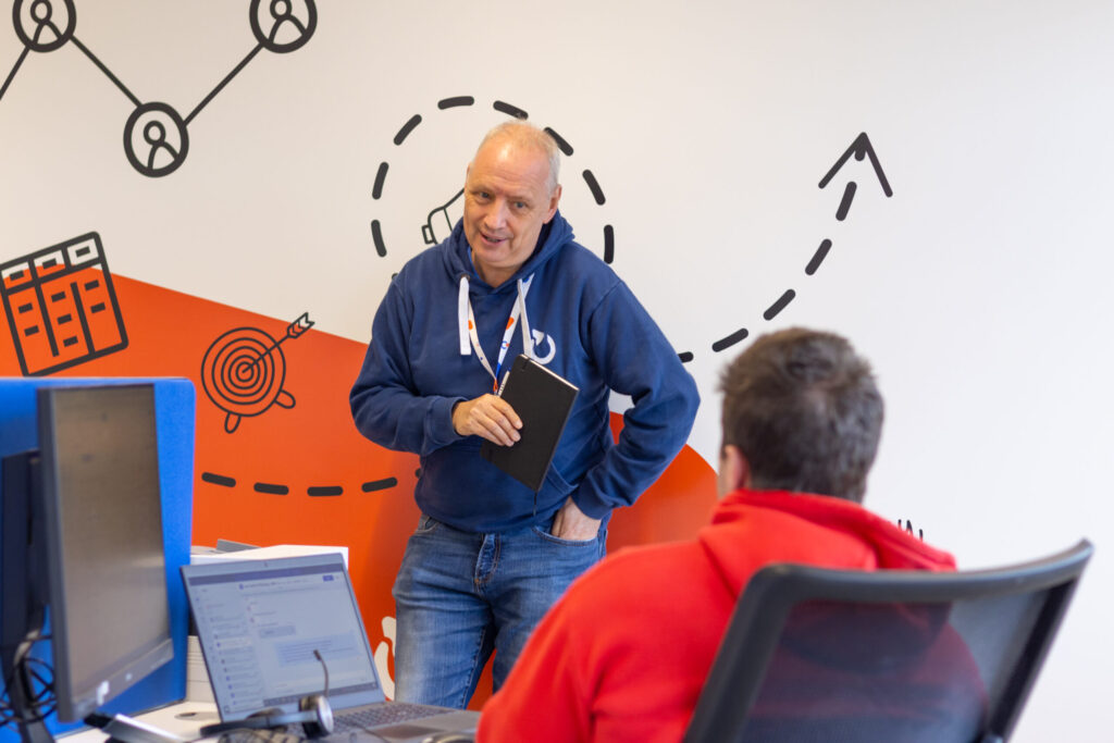 A man in the Spyrosoft office in Bournemouth leans against a wall covered with a colorful abstract mural. He looks at a man sitting in an office chair.