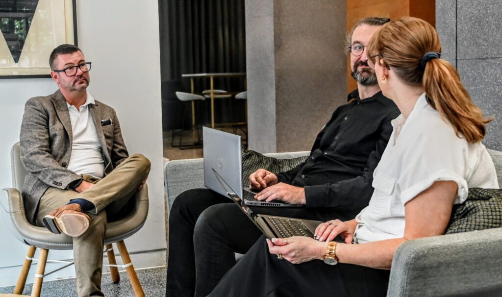 A man sits in an armchair, and a man and a woman sit on a couch next to him. They are working on a joint project at the Spyrosof site in London.