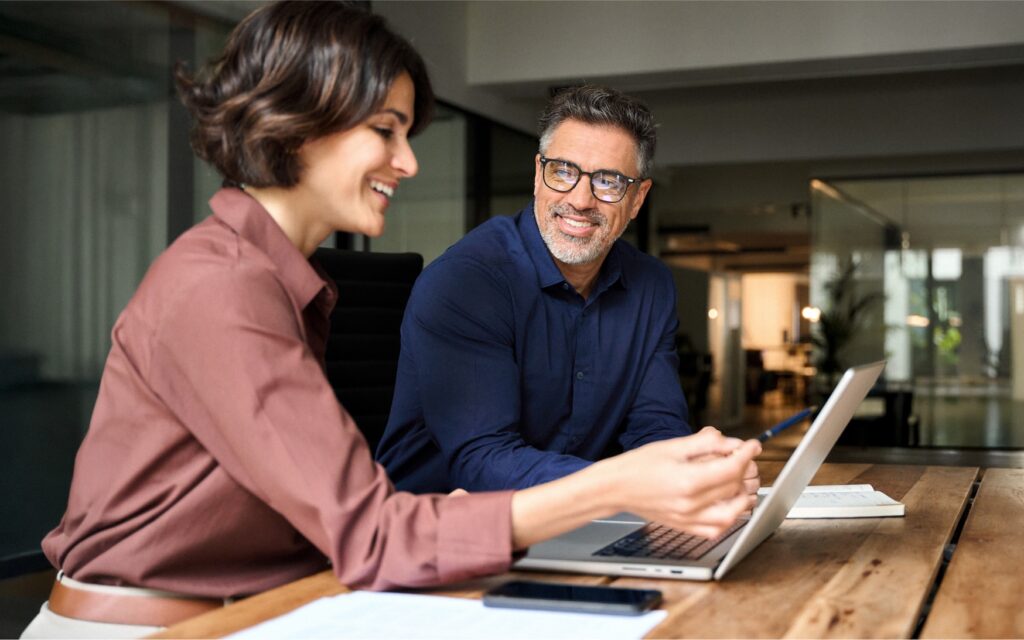 Two colleagues smiling and collaborating at a desk, reviewing information together on a laptop in a modern office.