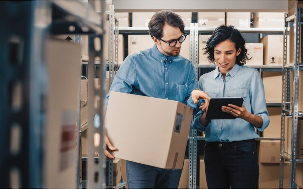 Two warehouse workers reviewing inventory – one holding a cardboard box while the other checks details on a tablet between storage shelves.