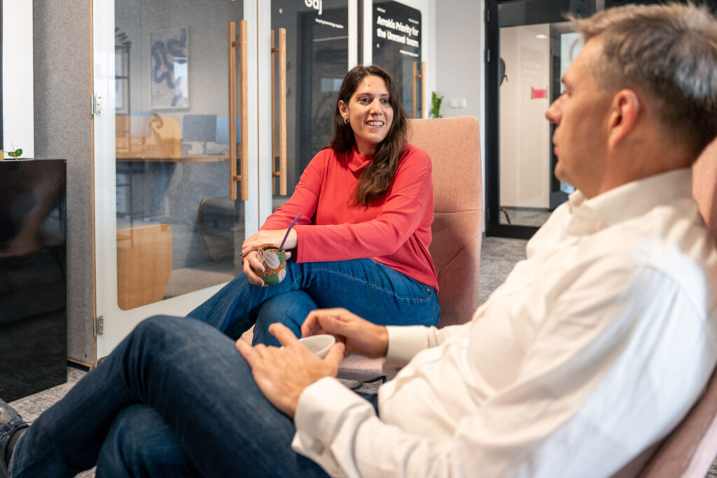 A woman in a red sweater talks to a man in a white shirt. They both sit in armchairs next to conference booths in Spyrosoft's Warsaw office.